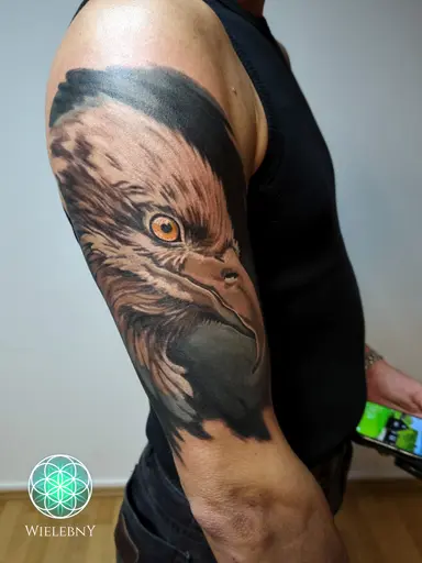 Realistic black and gray eagle head tattoo with a vivid orange eye on the upper outer arm and shoulder.